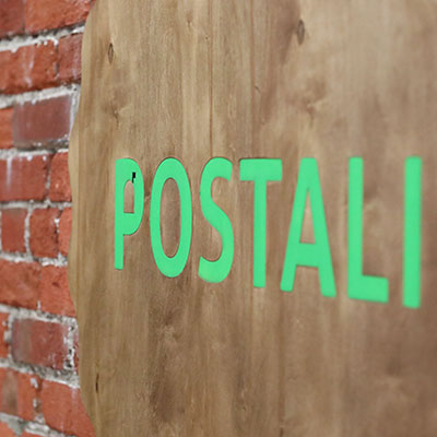 A wooden sign with the word POSTALI in bright green letters is mounted on a wall with exposed red bricks.