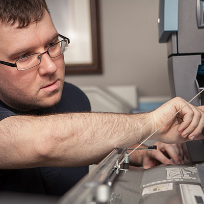 A man wearing glasses is repairing or adjusting a piece of office equipment, possibly a printer or copier, focusing intently as he works with a tool inside the machine.