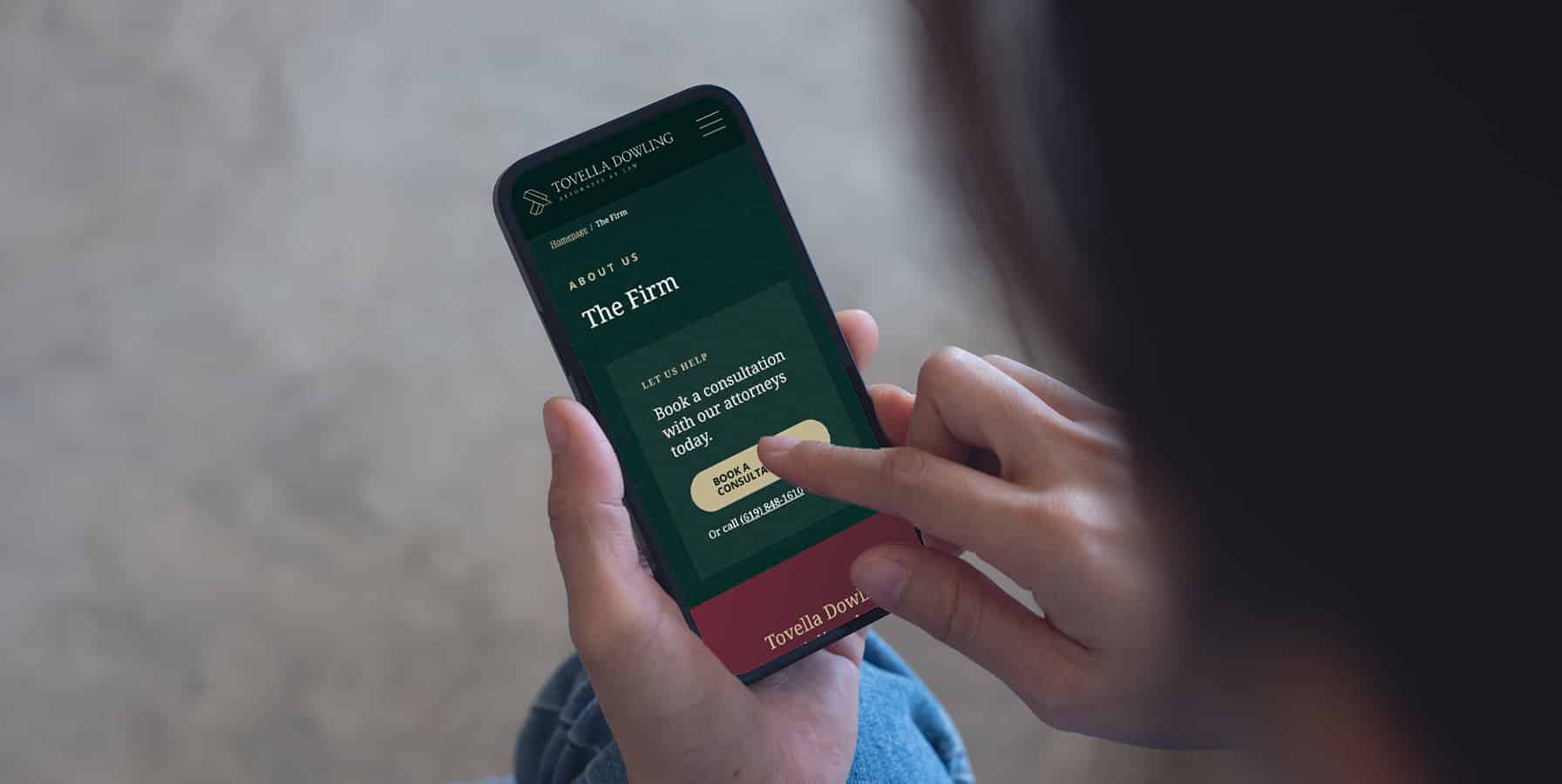 Person holding a smartphone and using their finger to interact with a law firms website, specifically on a page titled The Firm with a button to book a consultation. The screen background is dark green.