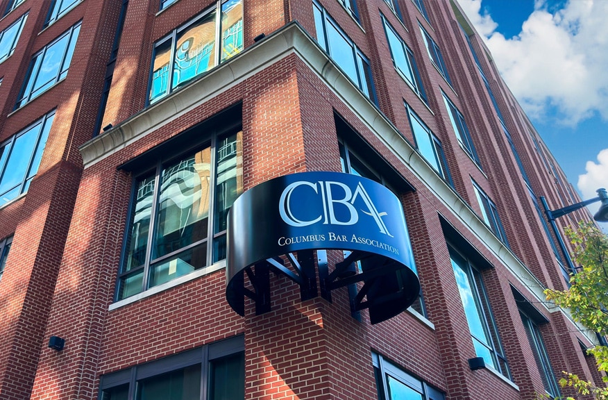 A brick building with a blue curved sign that reads CBA Columbus Bar Association above an entrance, with large windows and a partly cloudy sky in the background.