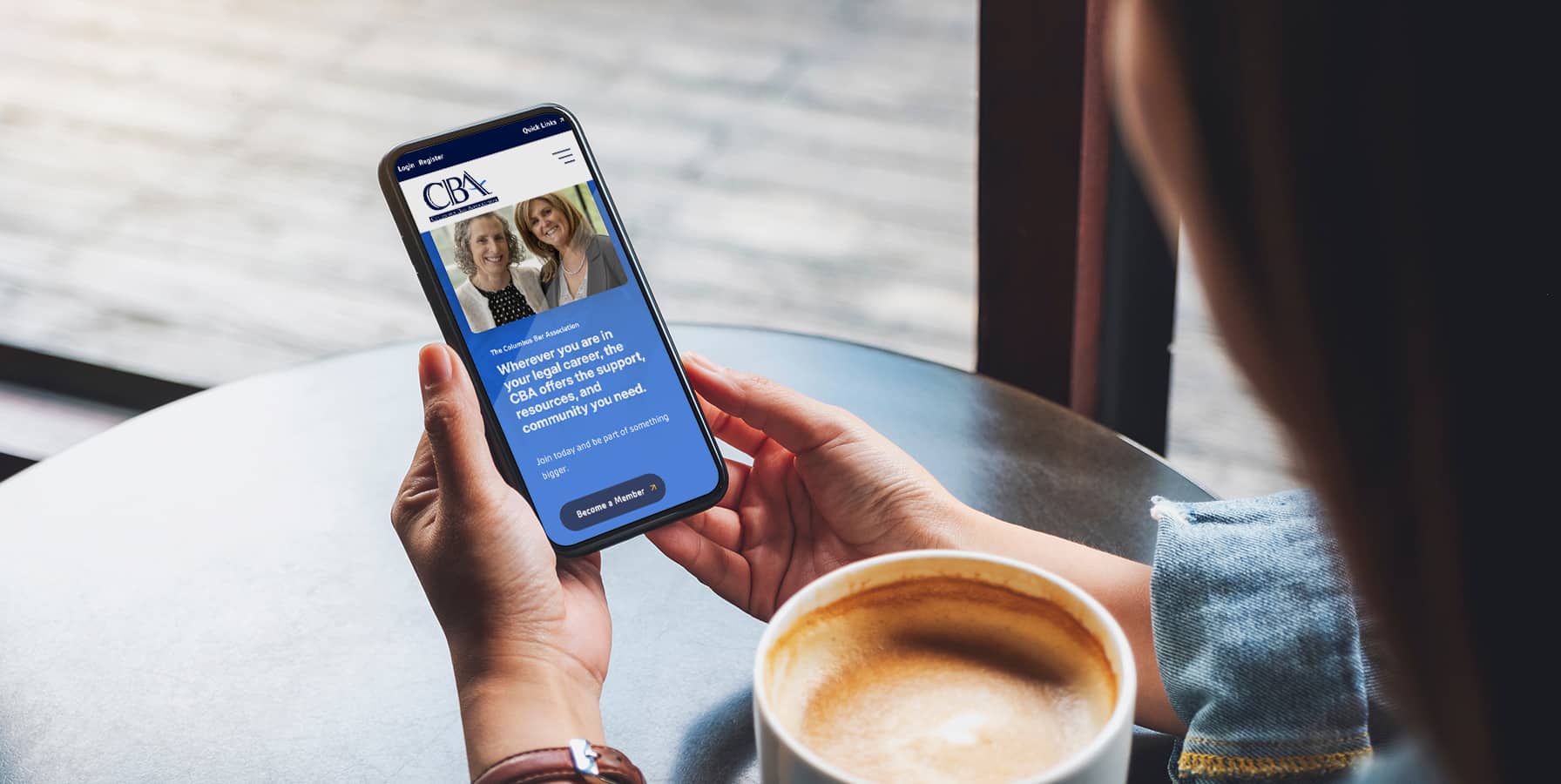 A person holding a smartphone at a café table with a cup of coffee, browsing a website featuring two women and information about consulting and business support.