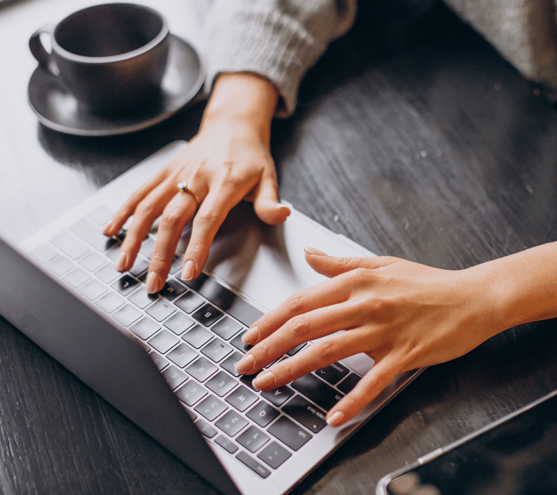 Close-up of a person typing on a laptop keyboard at a dark wooden table, with a cup and saucer and a smartphone nearby. The person is wearing a gray sweater and a ring.