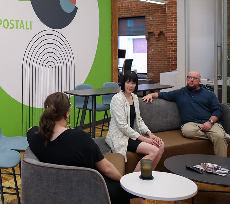 Three people are sitting and talking in a modern office lounge area with colorful wall art, a round table, and magazines on the table. The atmosphere is casual and relaxed.