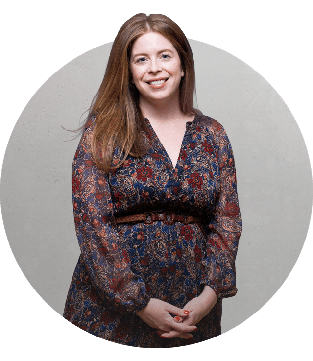 A woman with long brown hair, wearing a patterned dress with red and blue tones, stands smiling with her hands clasped in front of her against a light gray circular background.
