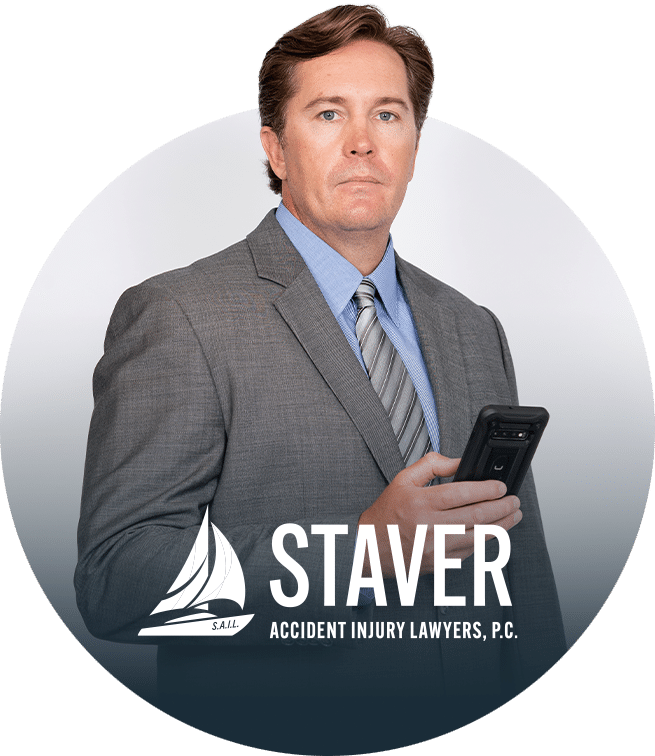 A man in a gray suit holding a smartphone looks at the camera; the logo and text Staver Accident Injury Lawyers, P.C. appear at the bottom of the image.